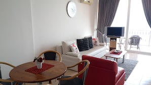 Un salon moderne avec un canapé blanc, un fauteuil rouge et une table à manger en bois. Un téléviseur à écran plat est fixé au mur, et il y a un balcon avec vue.