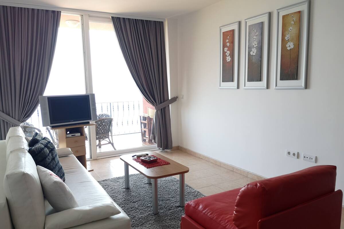 Een woonkamer met een bank, een rode stoel, een televisie en een balkon met gordijnen.