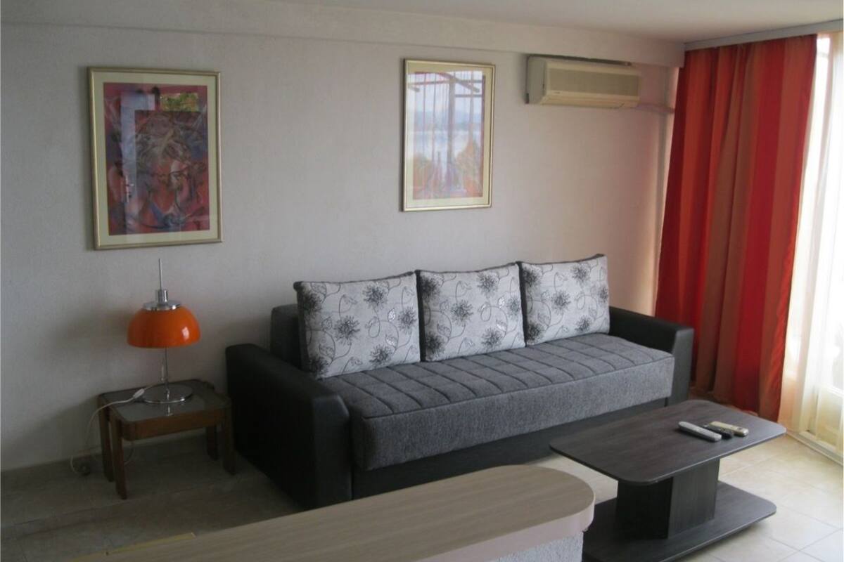 Een moderne woonkamer met een grijze bank, een klein bijzettafeltje met een oranje lamp en een salontafel. Er hangen twee ingelijste foto’s aan de muur en een raam met rode gordijnen.