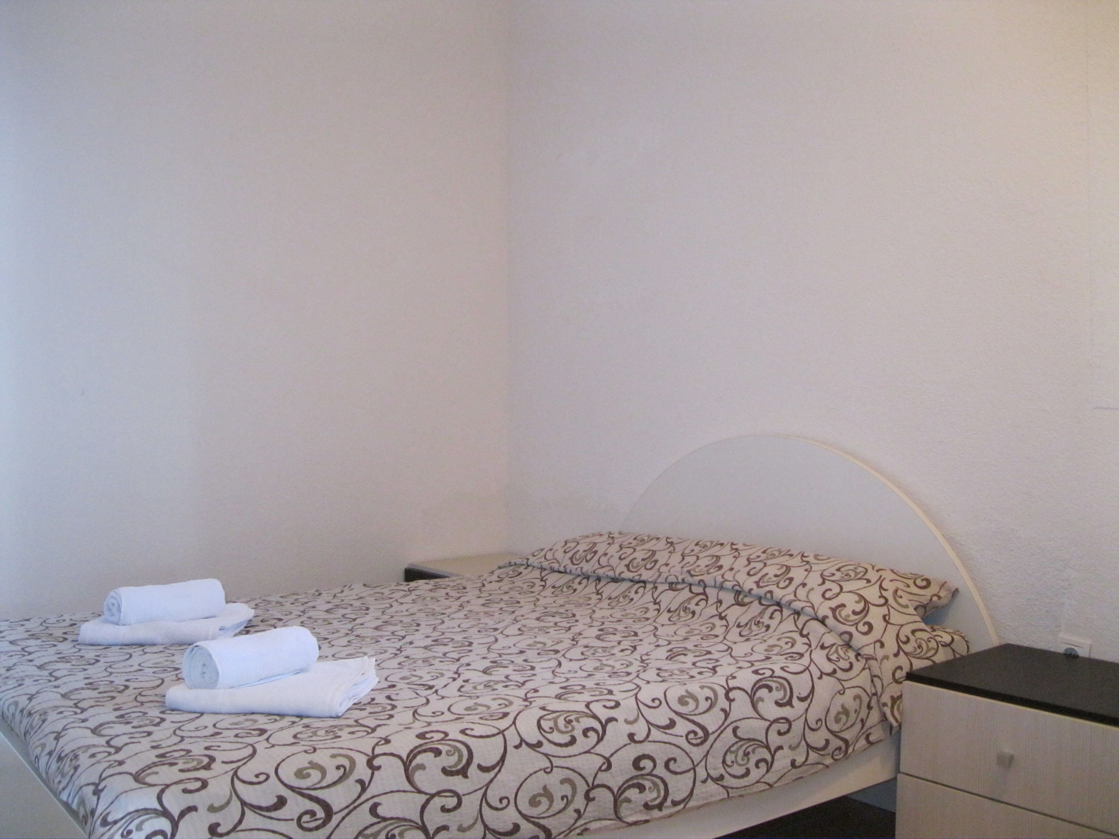 A single bed with a patterned bedspread and two rolled towels on it.