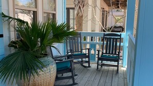 Une véranda en bois avec une grande plante en pot, deux fauteuils à bascule et un ventilateur de plafond.