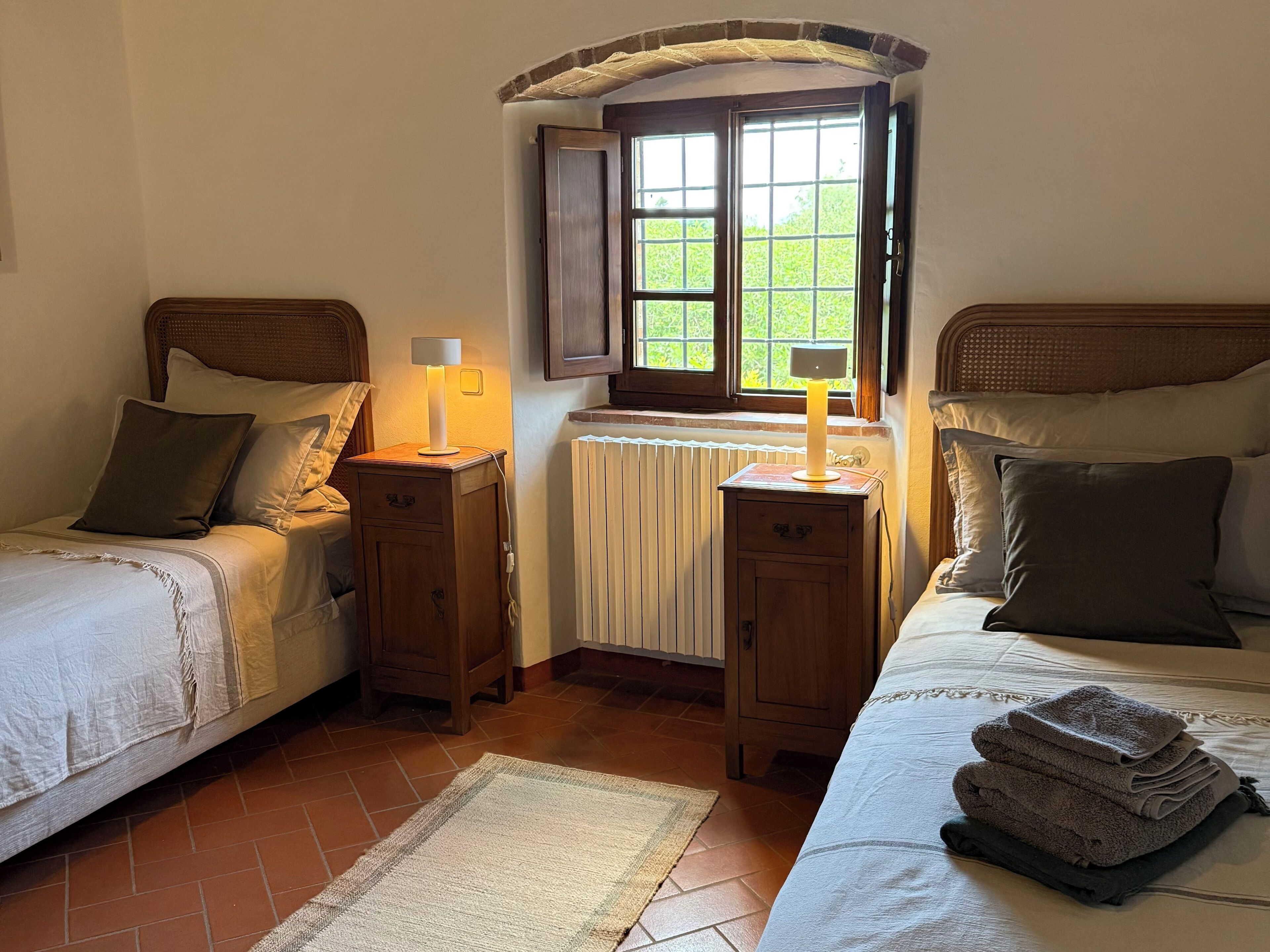 A bedroom with two beds, a window with a view of greenery, a radiator, and a bedside table with a lamp.