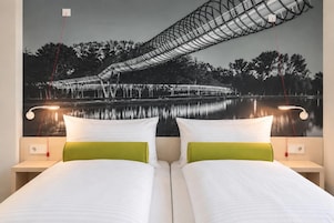 Una cama doble con ropa de cama blanca y verde, dos mesitas de noche con lámparas y un mural en la pared que muestra un puente sobre un cuerpo de agua.