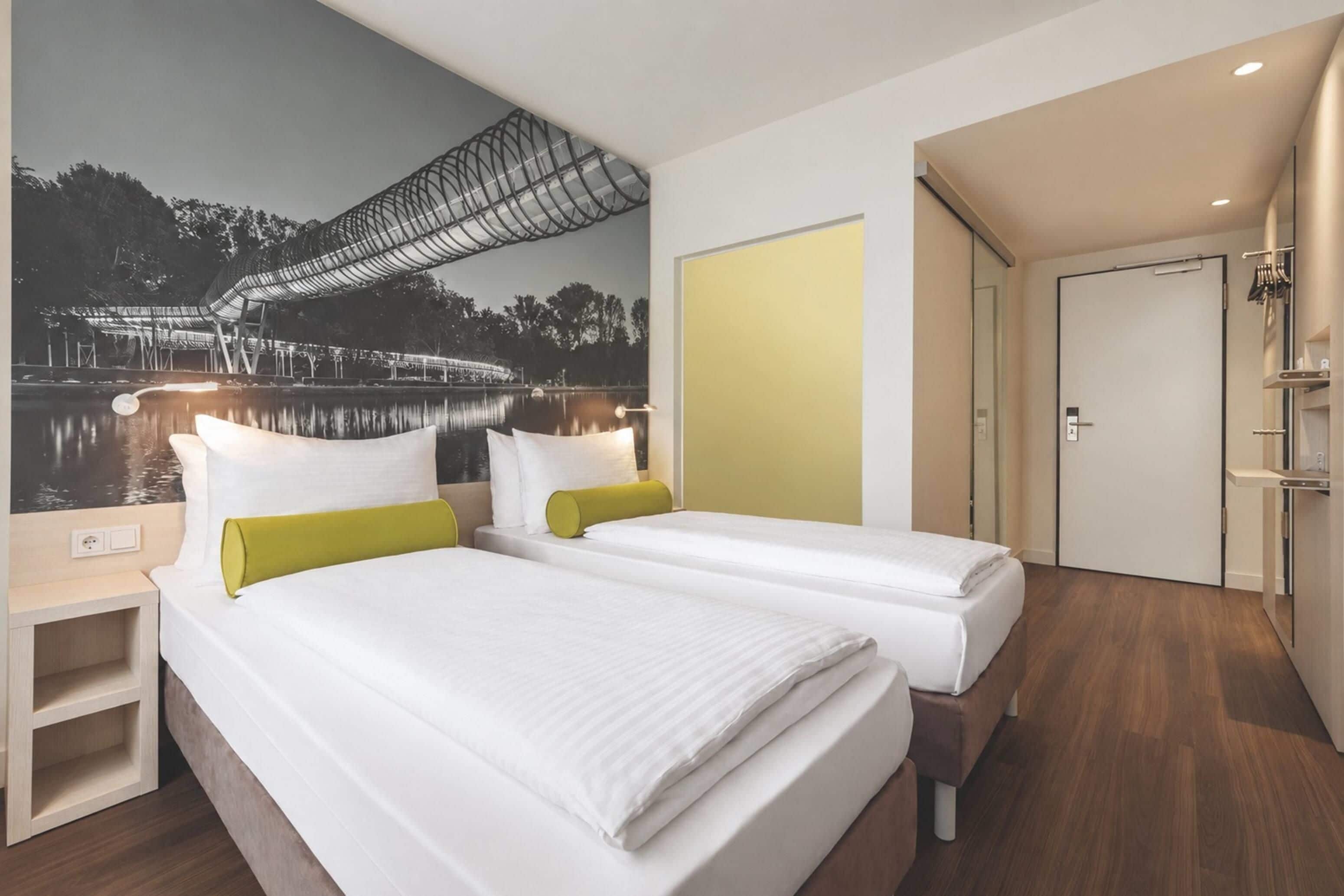 Ein Hotelzimmer mit zwei Betten, einem Wandbild einer Brücke über Wasser und einer Tür zum Badezimmer.