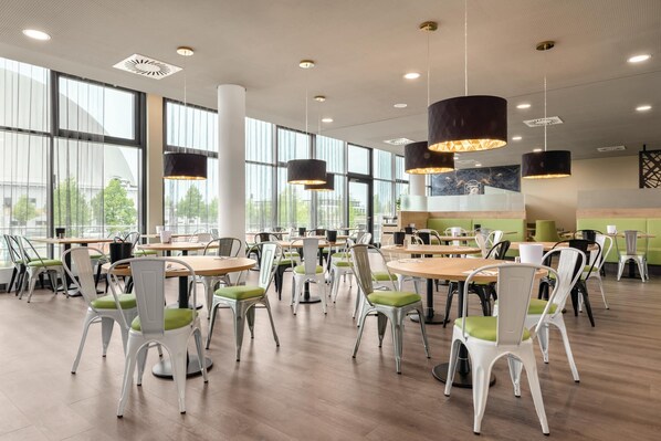 A spacious cafeteria with round tables and chairs, large windows, and pendant lighting. - Super 8 by Wyndham Oberhausen am Centro (Oberhausen)