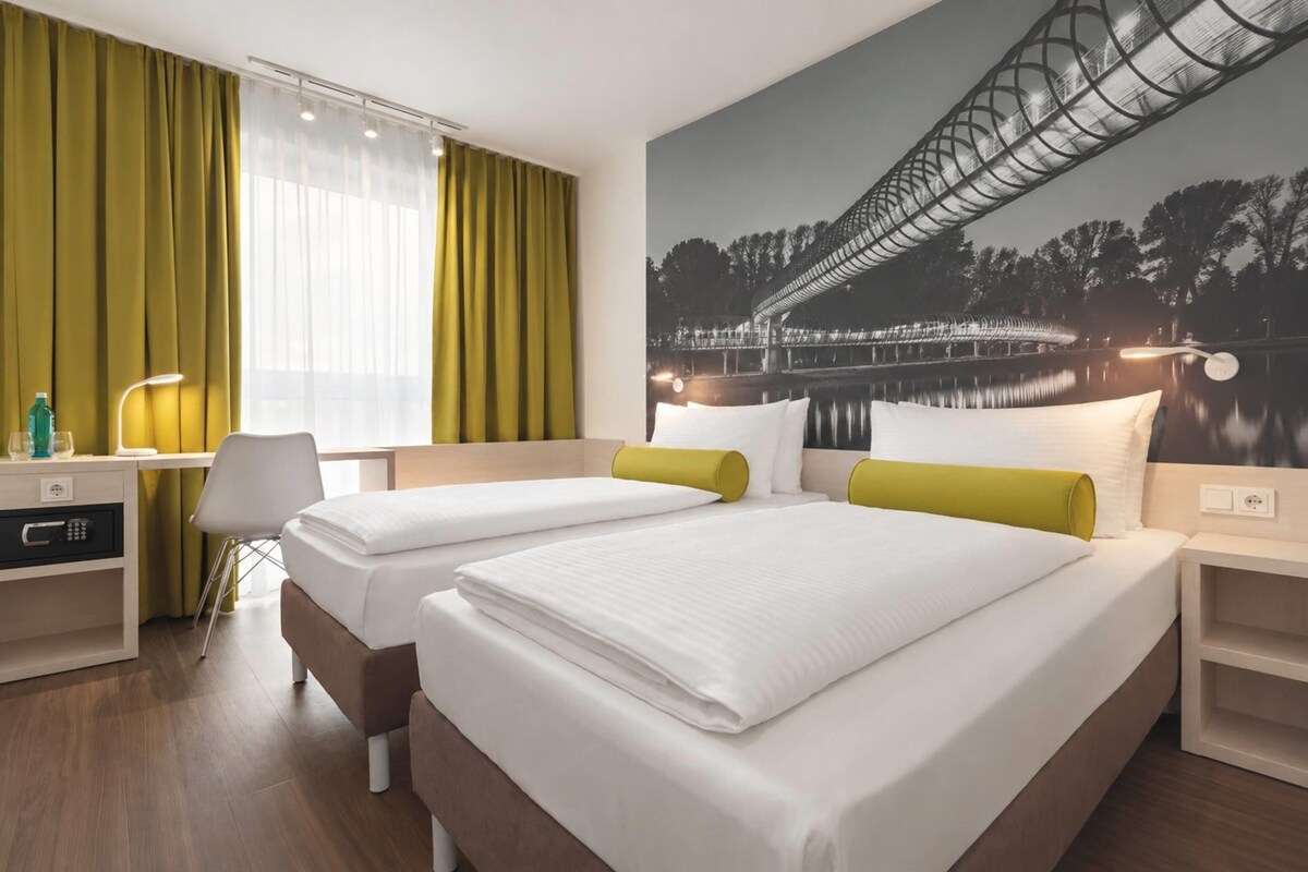 A hotel room with two beds, a desk, a chair, and a large window with curtains.