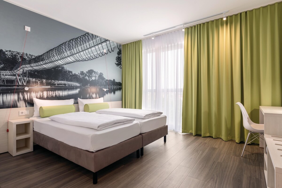 Een hotelkamer met een bed, nachtkastjes, een bureau, een stoel en een groot raam met gordijnen.