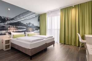 Ein Hotelzimmer mit Bett, Nachttischen, Schreibtisch, Stuhl und einem großen Fenster mit Vorhängen.