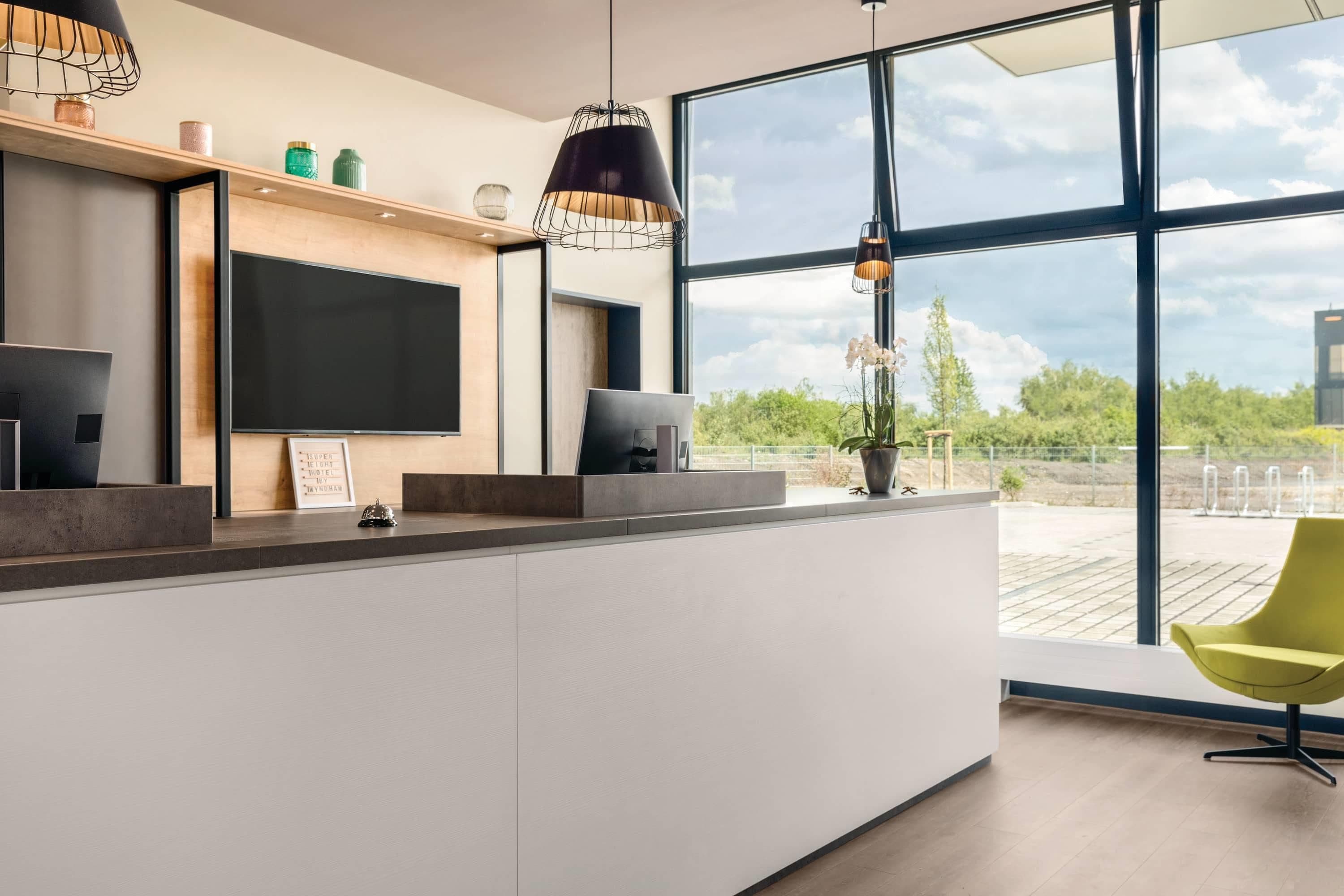 A modern hotel reception area with a large TV screen, two pendant lights, and a view of the outdoors through large windows.