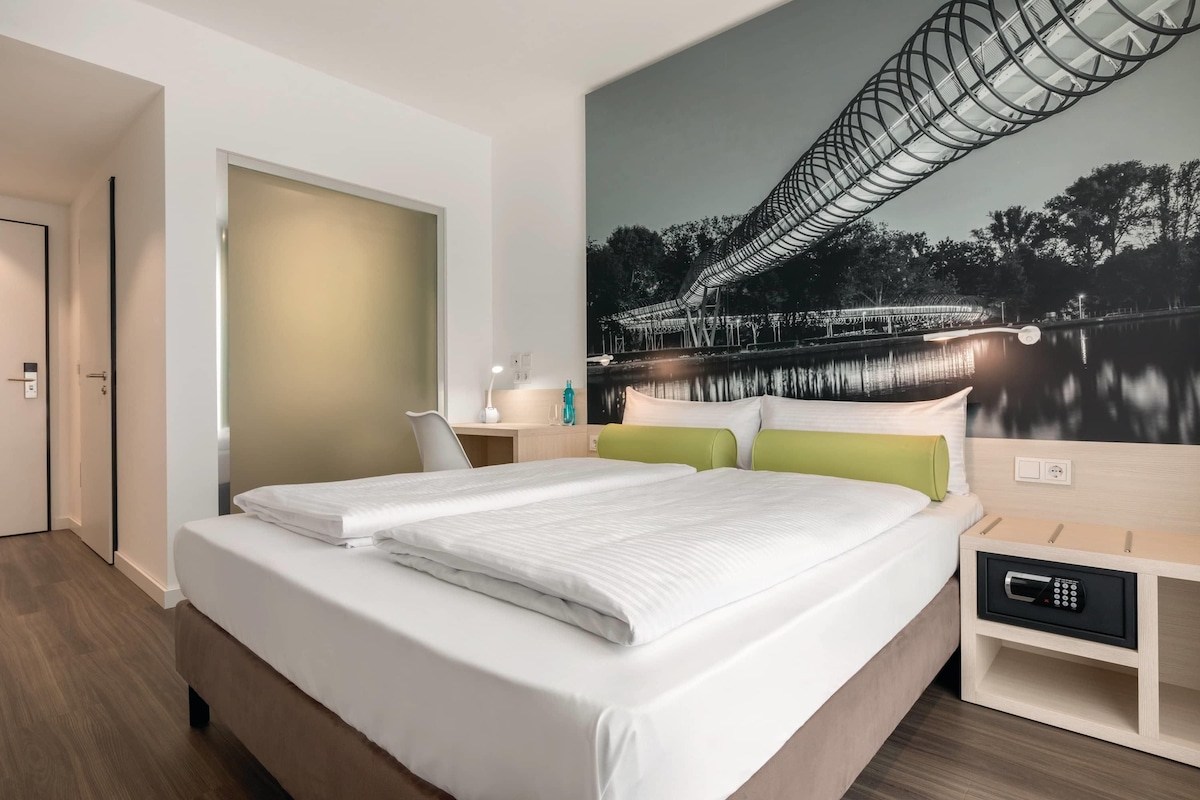 A hotel room with a large bed, a desk, a chair, and a wall-mounted TV.