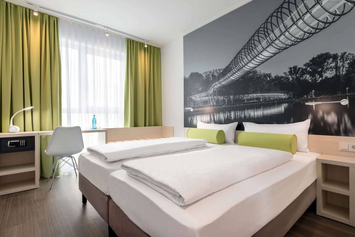 Una habitación de hotel con dos camas, un escritorio, una silla y un ventanal con cortinas.