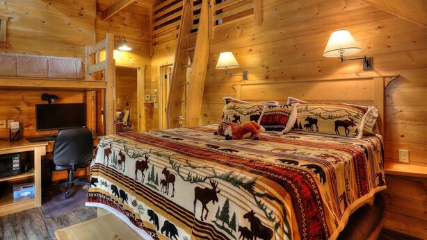 Ein gemütliches Blockhauszimmer mit einem Bett mit tierischen Motiven, einem Schreibtisch mit Computer und zwei Wandleuchten.