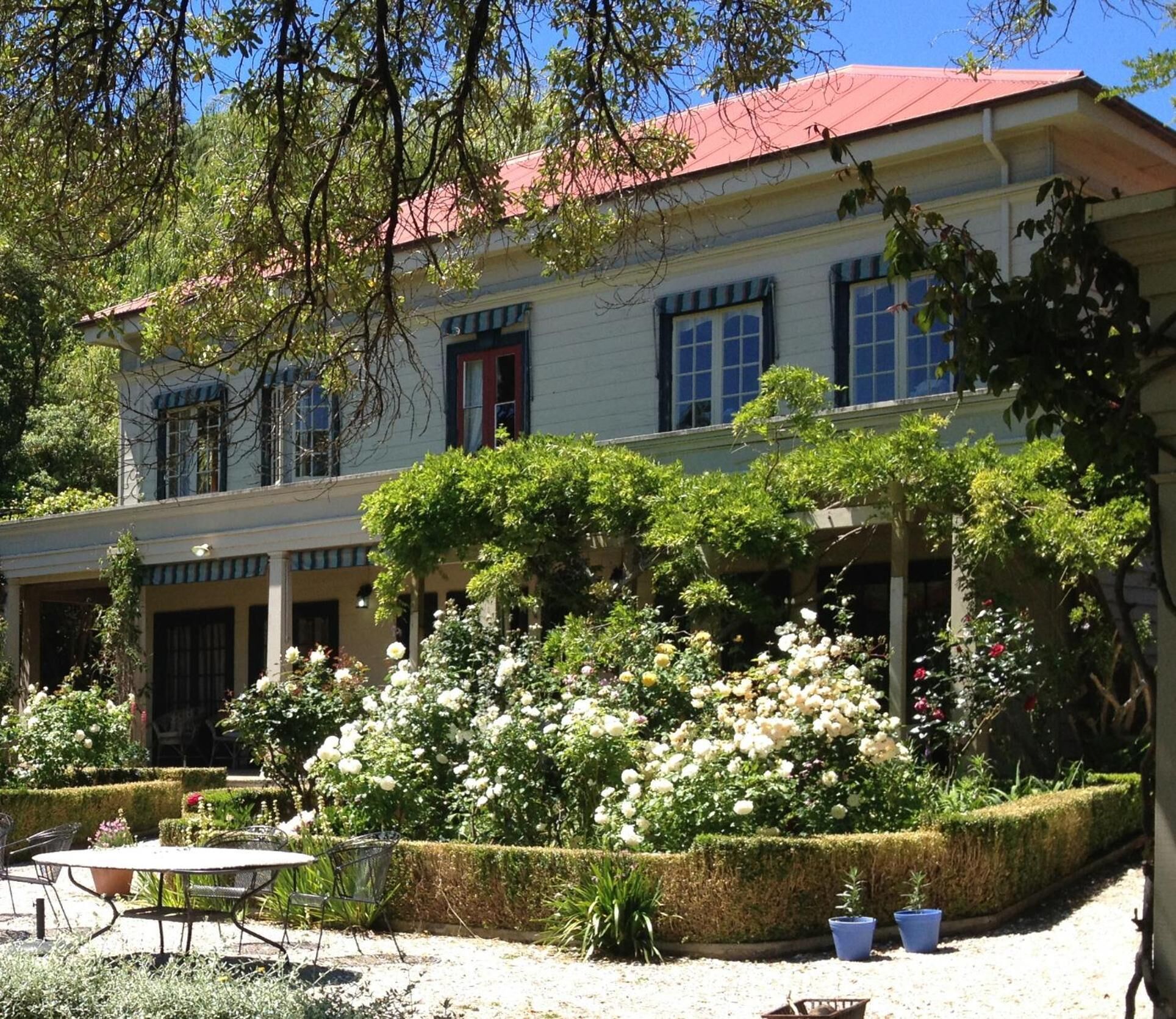 A two-story building with a red roof, surrounded by lush greenery and flowering plants.