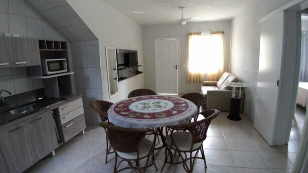 A compact kitchen with a microwave, sink, and cabinets. There is a dining area with wicker chairs and a table. A sofa and a lamp are visible in the background. - Residencial Malu (Penha)