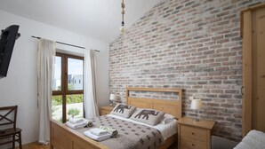A bedroom with a brick wall, a wooden bed, a window with curtains, a bedside table with a lamp, and a chair. - Villa Paradis 6 - family villa with Pool (Butkovici)