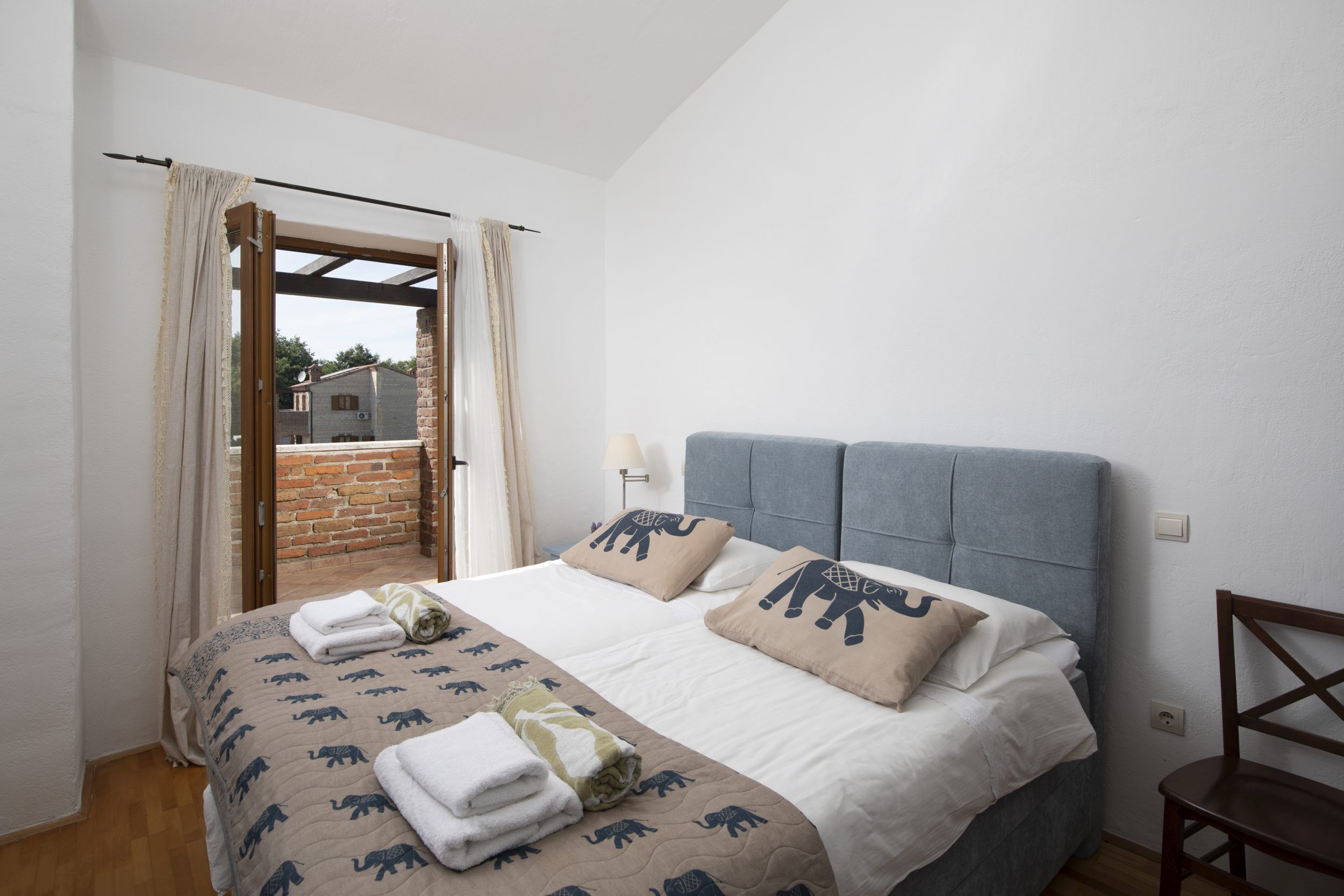 Une chambre à coucher avec un lit, une fenêtre avec des rideaux, une vue sur un bâtiment en briques à l’extérieur et une chaise en bois.