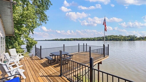 A wooden deck with lounge chairs and a table, overlooking a calm body of water with a flag. - Arkansas River House at the Foot Hills of Petit Jean Mt. (Morrilton)