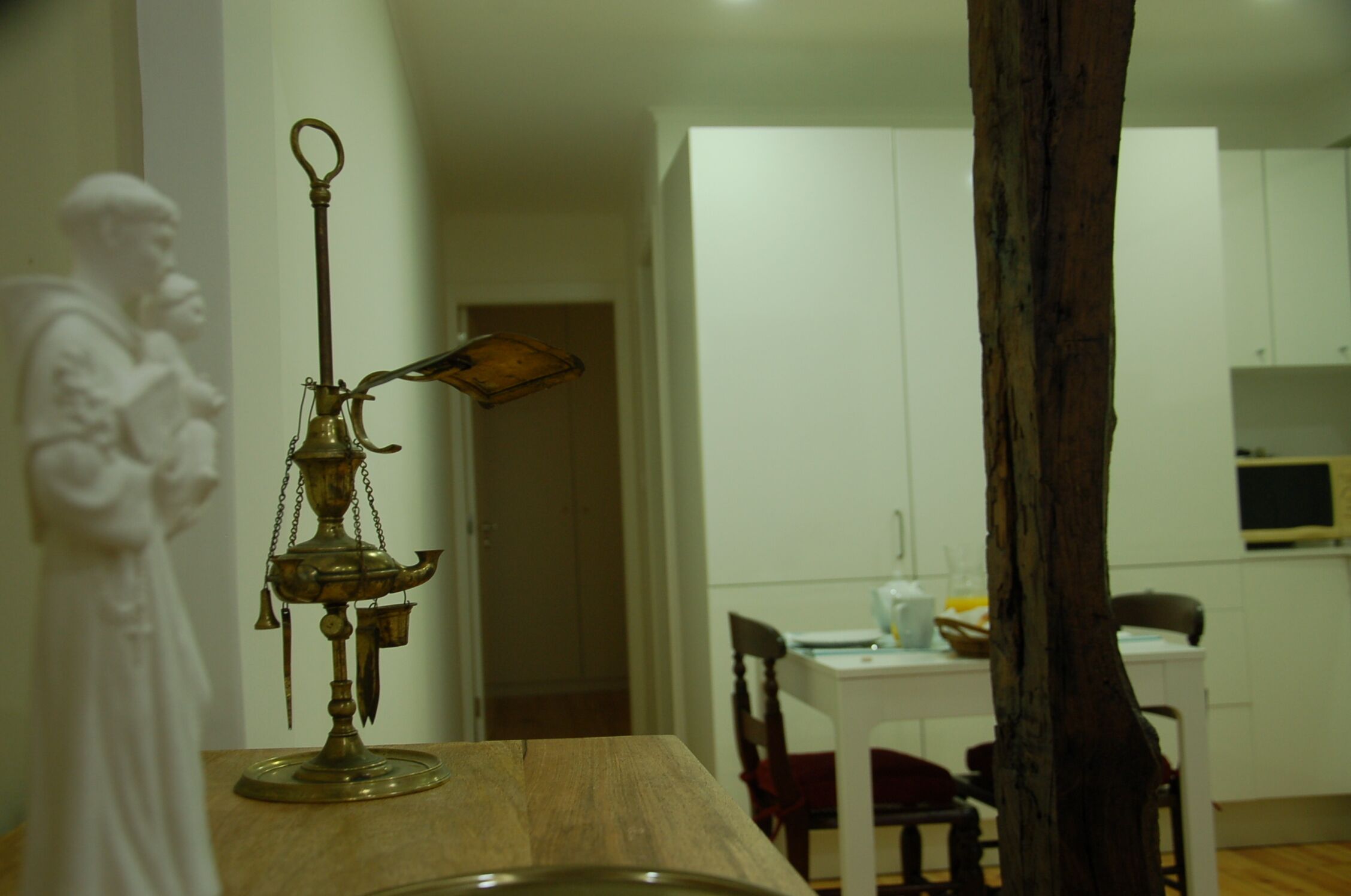 A dining area with a wooden table, chairs, and a brass lamp. There is a white statue on the left and a white cabinet in the background.
