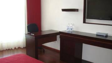 A hotel room with a wooden desk, a chair, a bed with a red blanket, a flat-screen TV mounted on the wall, and a small shelf with water bottles.