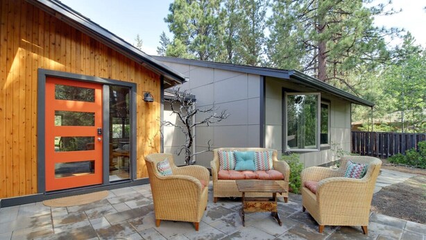 Une maison moderne avec une façade en bois, une porte d’un orange vif et un patio aménagé avec des meubles en osier et une petite table.