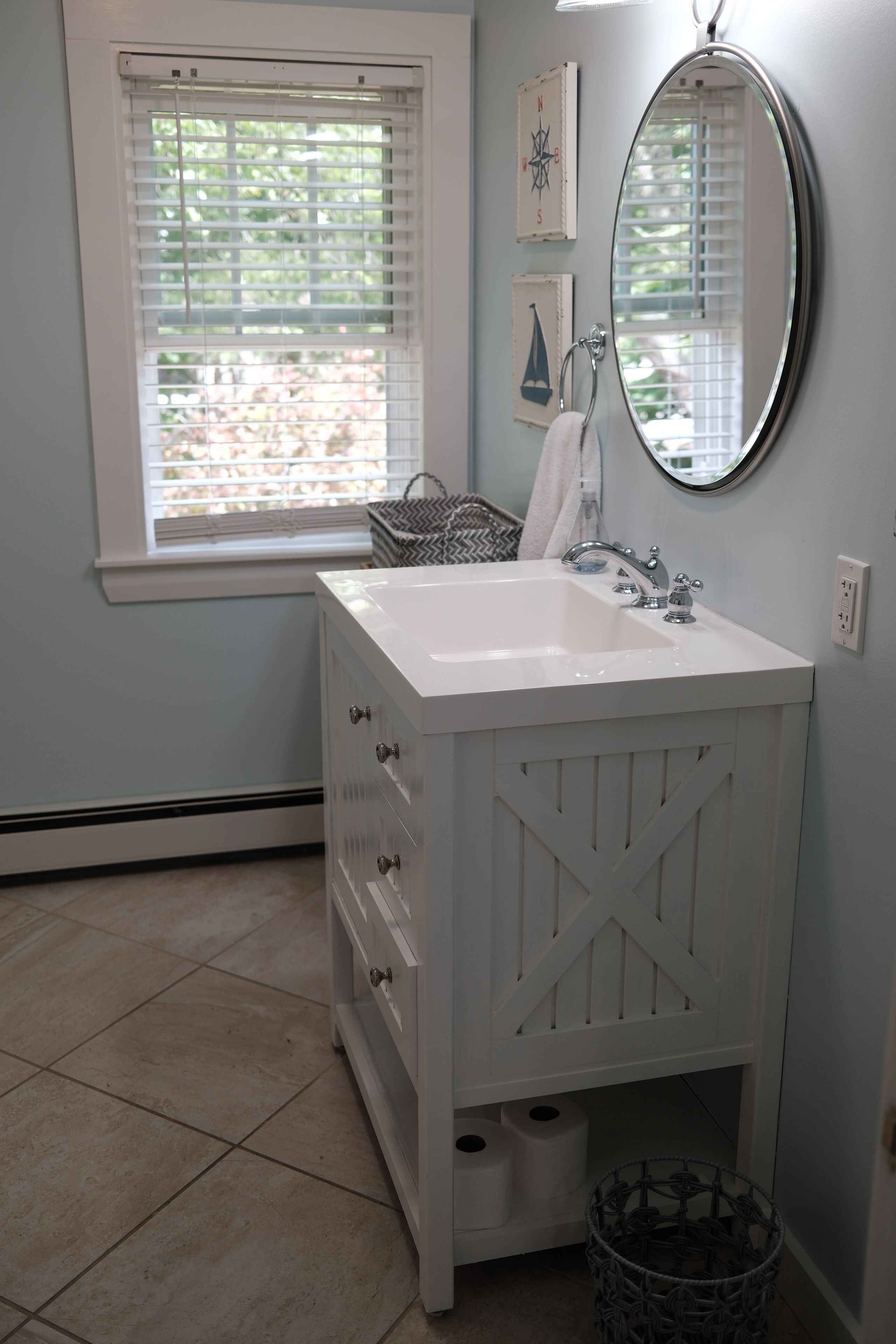 A bathroom with a white vanity, a round mirror, and a window with blinds.