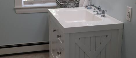 A bathroom with a white vanity, a round mirror, and a window with blinds.