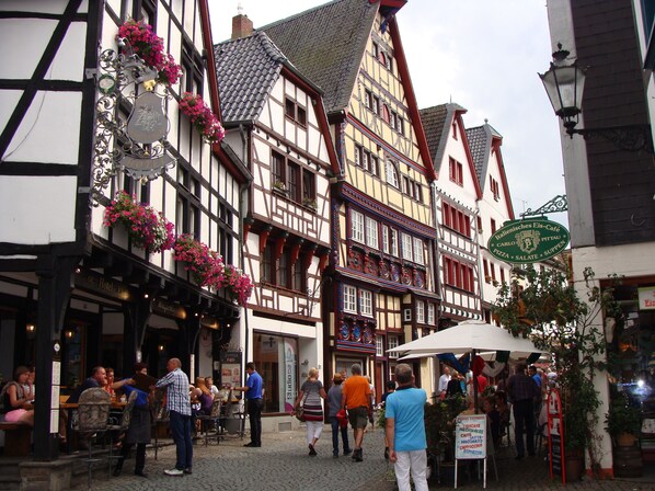 A traditional European street with half-timbered buildings, outdoor seating for cafes, and a sign for a local eatery.