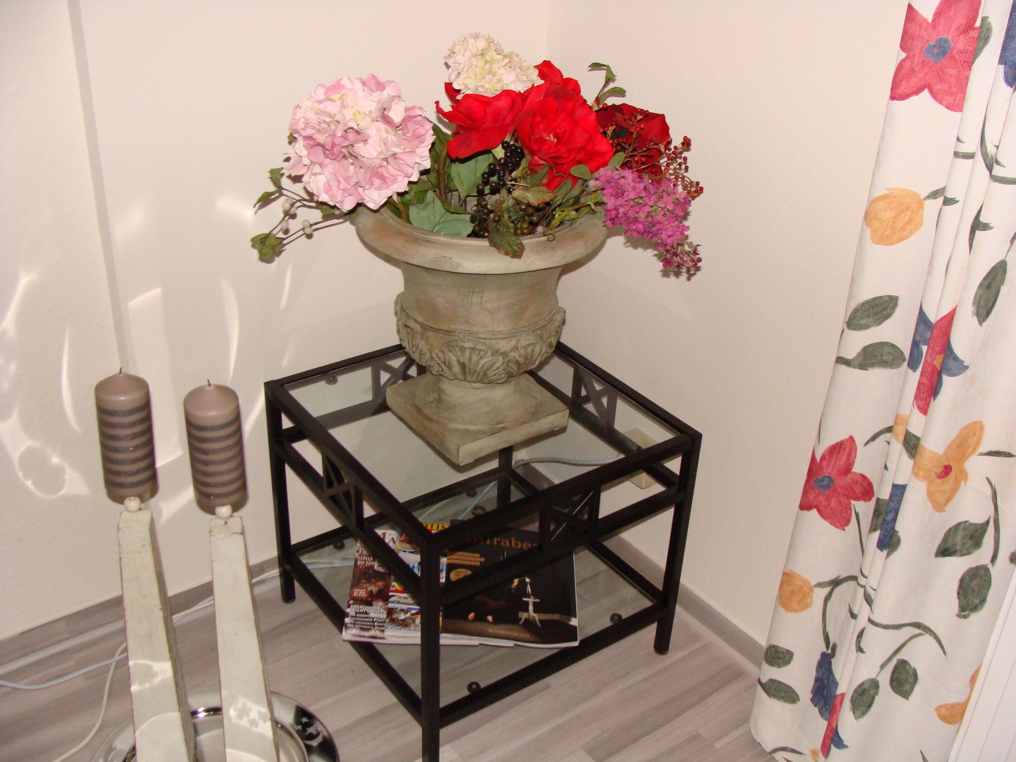 Ein Zimmer mit Glastisch, der eine Blumenvase, eine Kerze und Zeitschriften trägt.