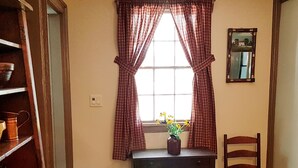 Ein Zimmer mit Fenster, einem Tisch mit Blumenvase, einem Stuhl und einem Regal mit Küchenutensilien.