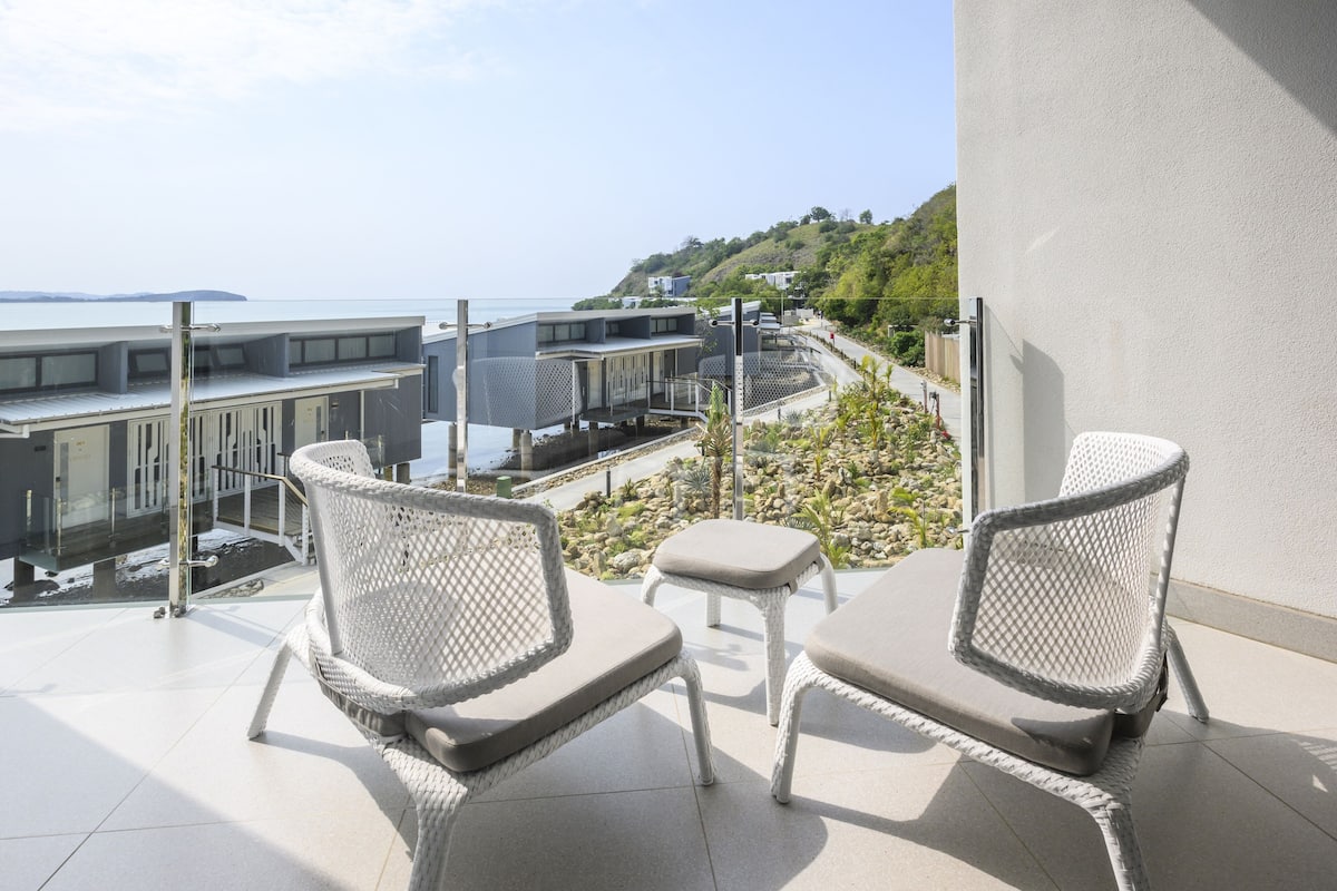 Un balcon doté de chaises en osier et d’une petite table, offrant une vue sur une zone côtière avec des bâtiments et sur la mer.
