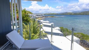 Un balcon doté de chaises longues blanches, d’une balustrade en verre et offrant une vue sur une mer calme et des collines lointaines.