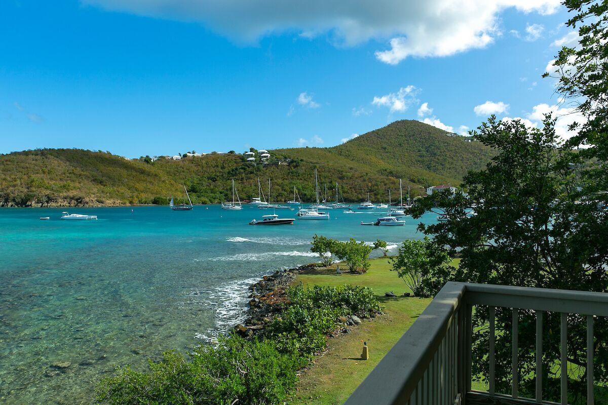 A coastal view with boats anchored in clear turquoise waters, a grassy area with a railing, and hills in the background.