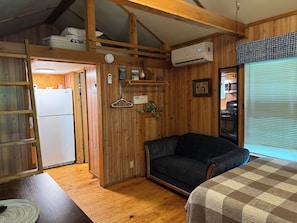 A cozy room with a bunk bed, a sofa, a refrigerator, an air conditioning unit, and a window with blinds.