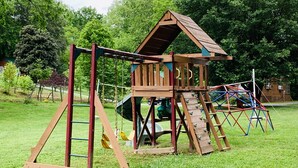 A wooden play structure with a climbing wall, a slide, and swings in a grassy area. - CREEKSIDE CABINS  - CHIPMUNK (Bryson City)