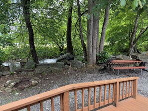 A wooden deck with picnic tables and benches, surrounded by trees and a rocky stream. - CREEKSIDE CABINS  - CHIPMUNK (Bryson City)