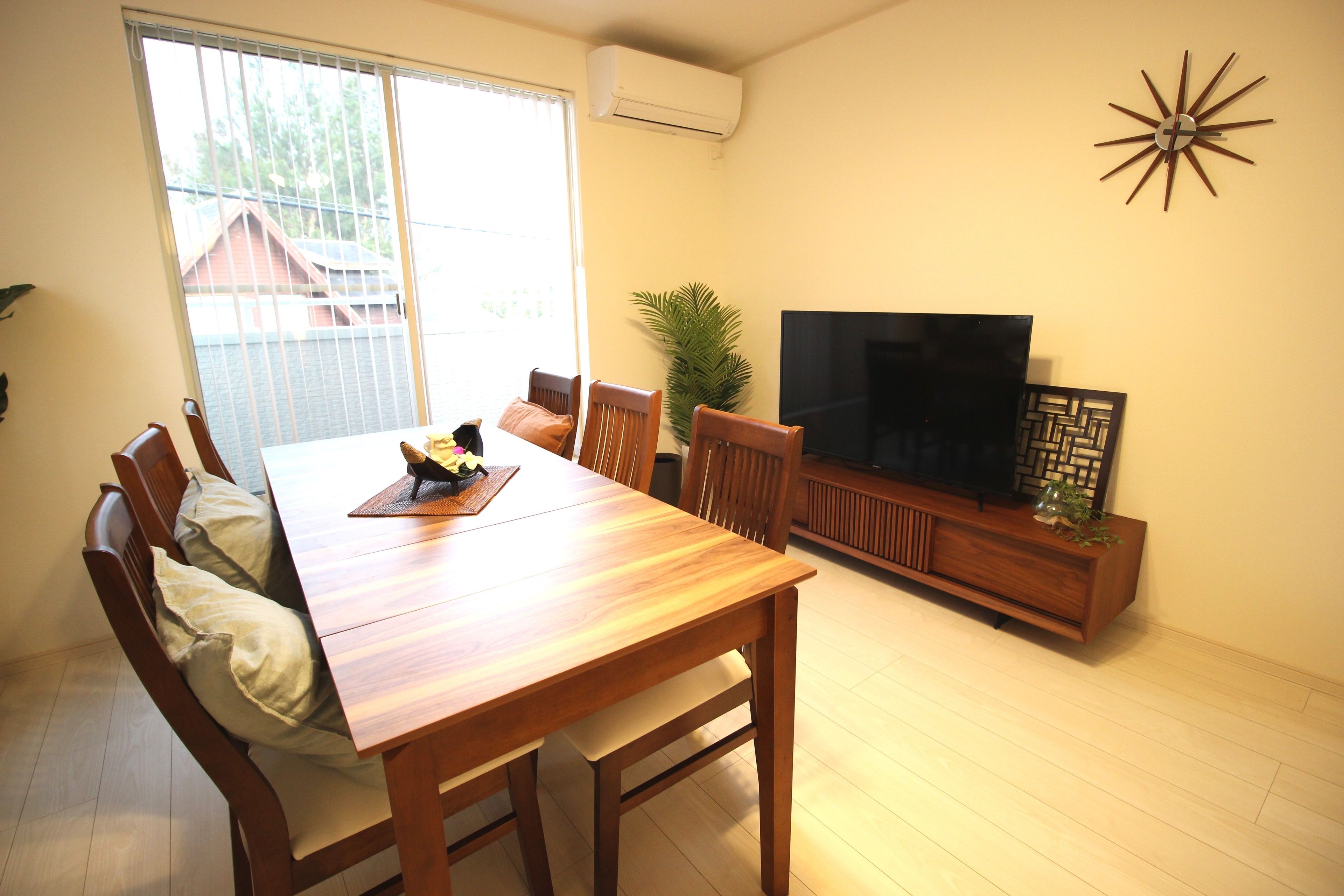 A dining area with a wooden table and chairs, a television on a wooden stand, and a large window with blinds.