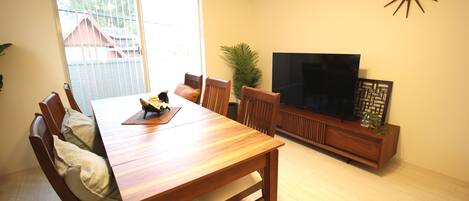 A dining area with a wooden table and chairs, a television on a wooden stand, and a large window with blinds.