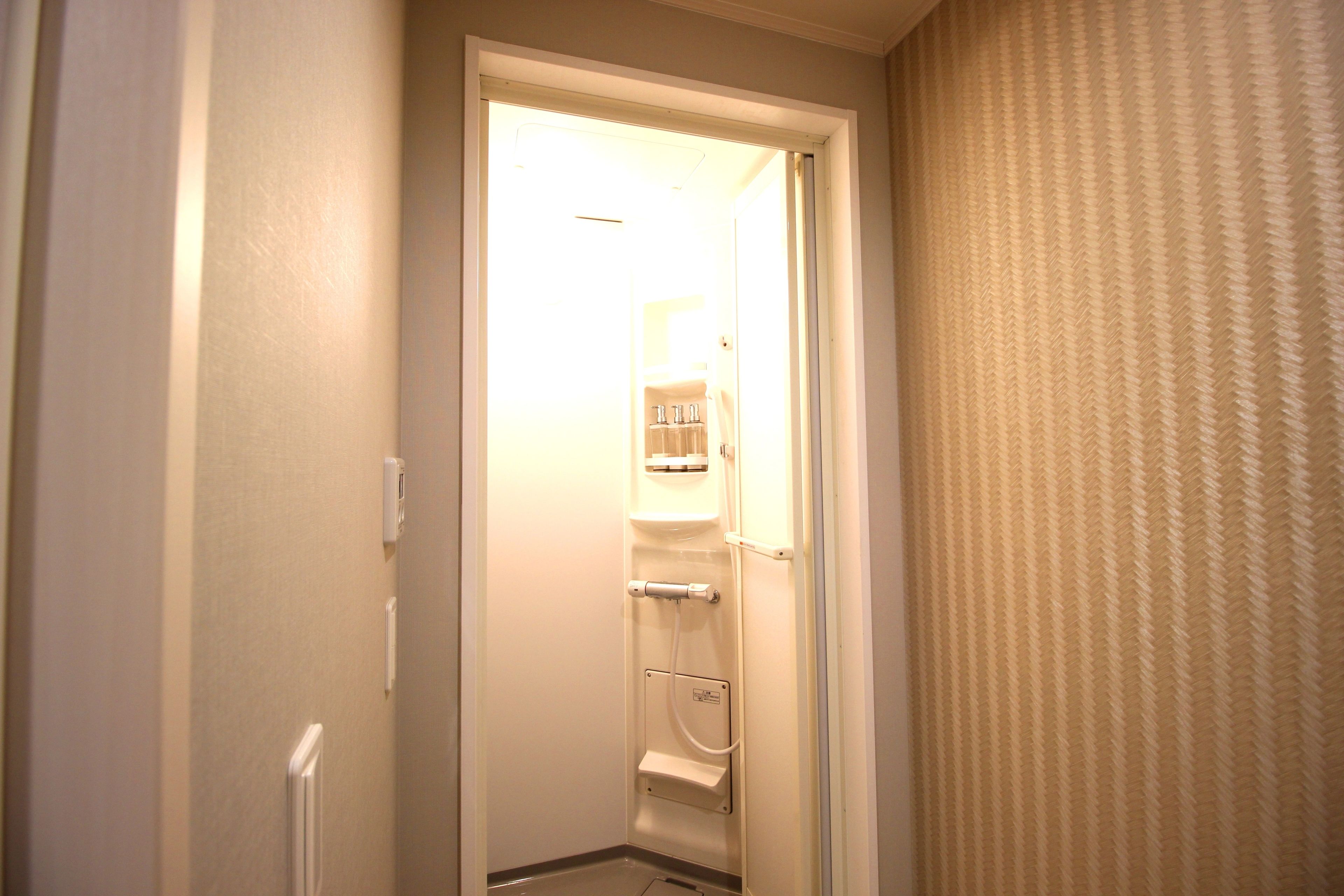 A narrow hallway with a door leading to a bathroom. The bathroom has a toilet, sink, and towel rack. The walls are plain, and there is a patterned curtain on the right.