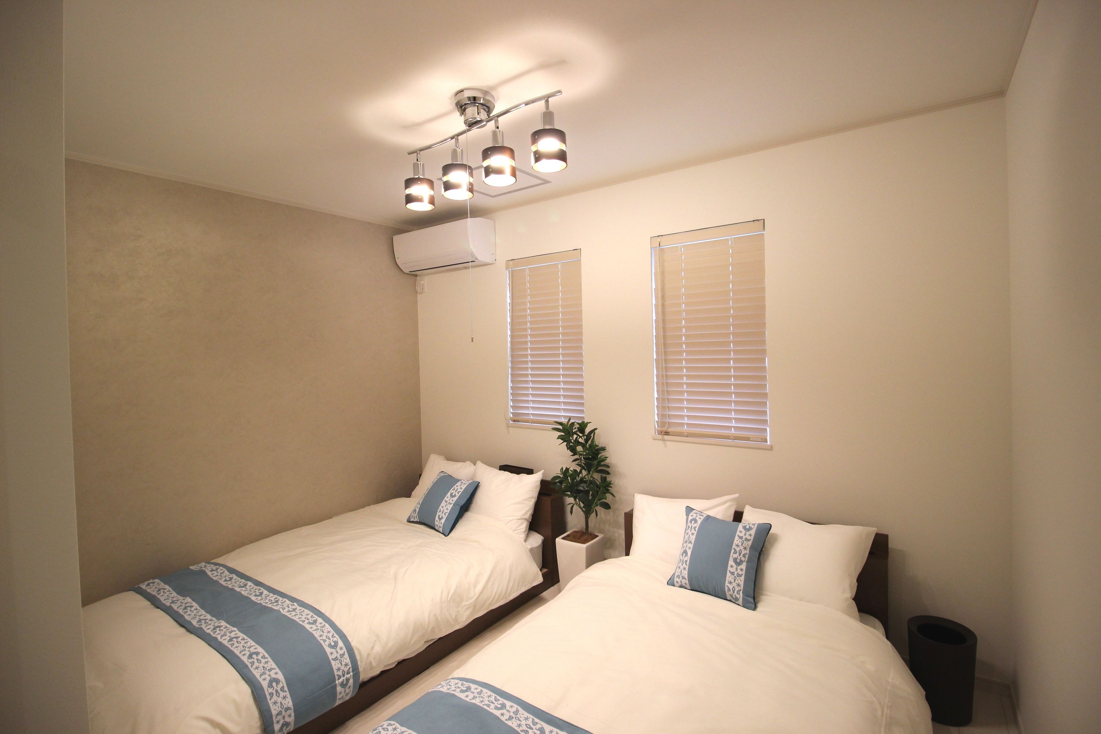 A bedroom with two beds, a ceiling light fixture, and two windows with blinds.