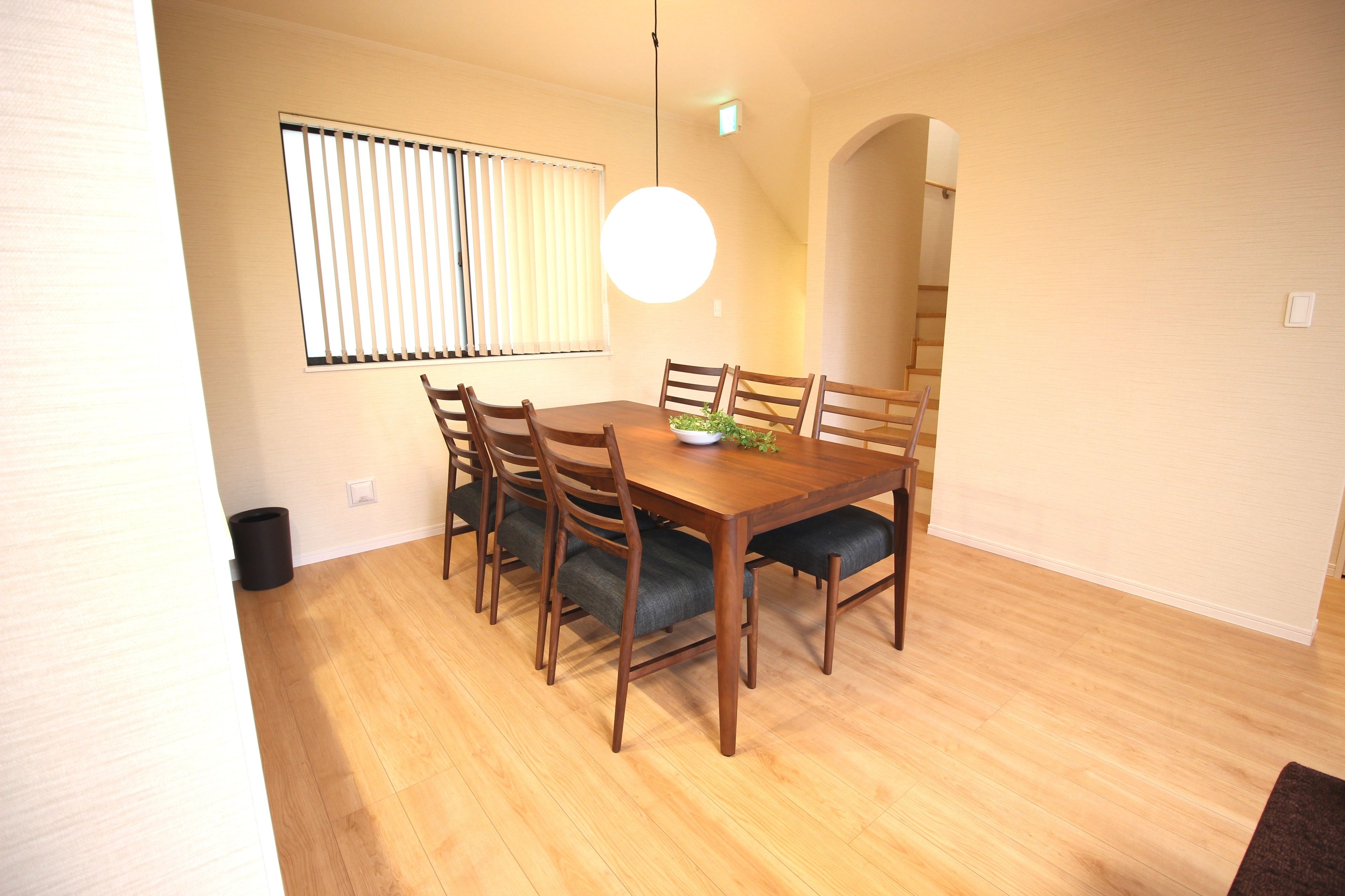 A dining area with a wooden table and chairs, a hanging light fixture, and a window with blinds.