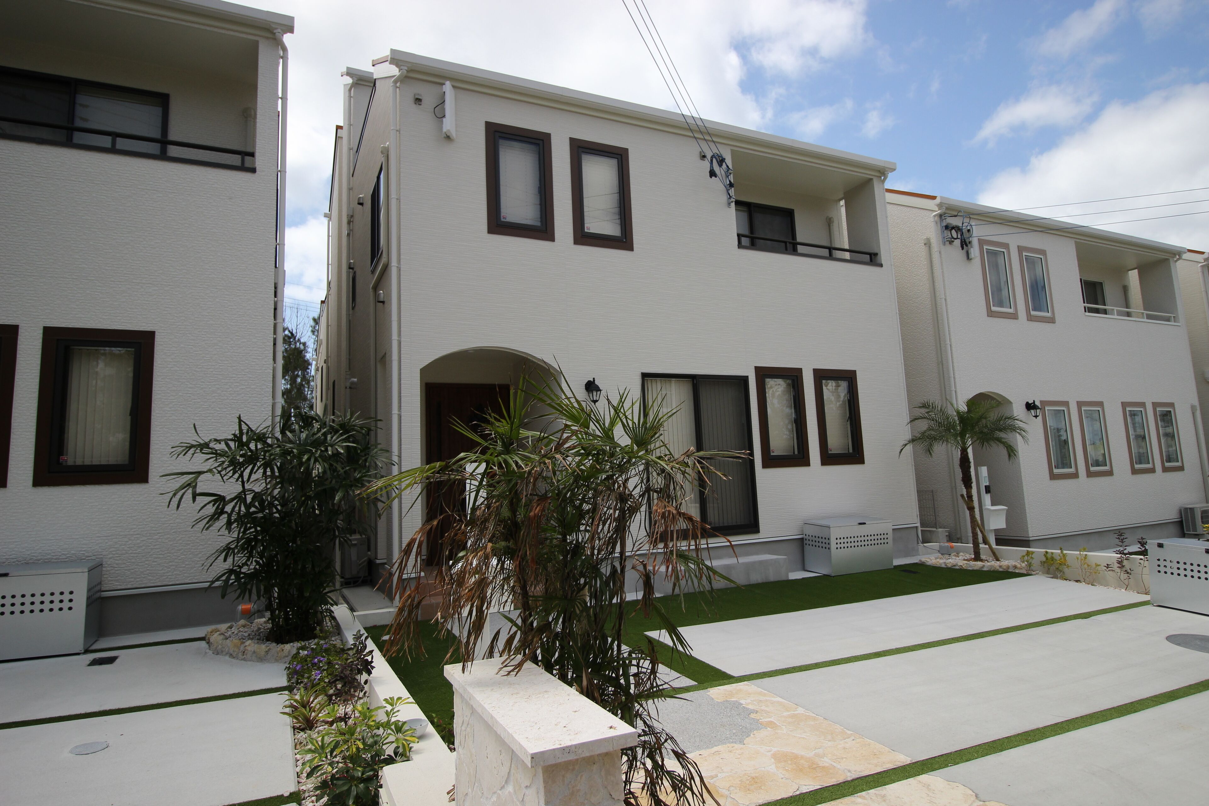A two-story building with a flat roof, multiple windows, and a small garden area with plants and a palm tree.