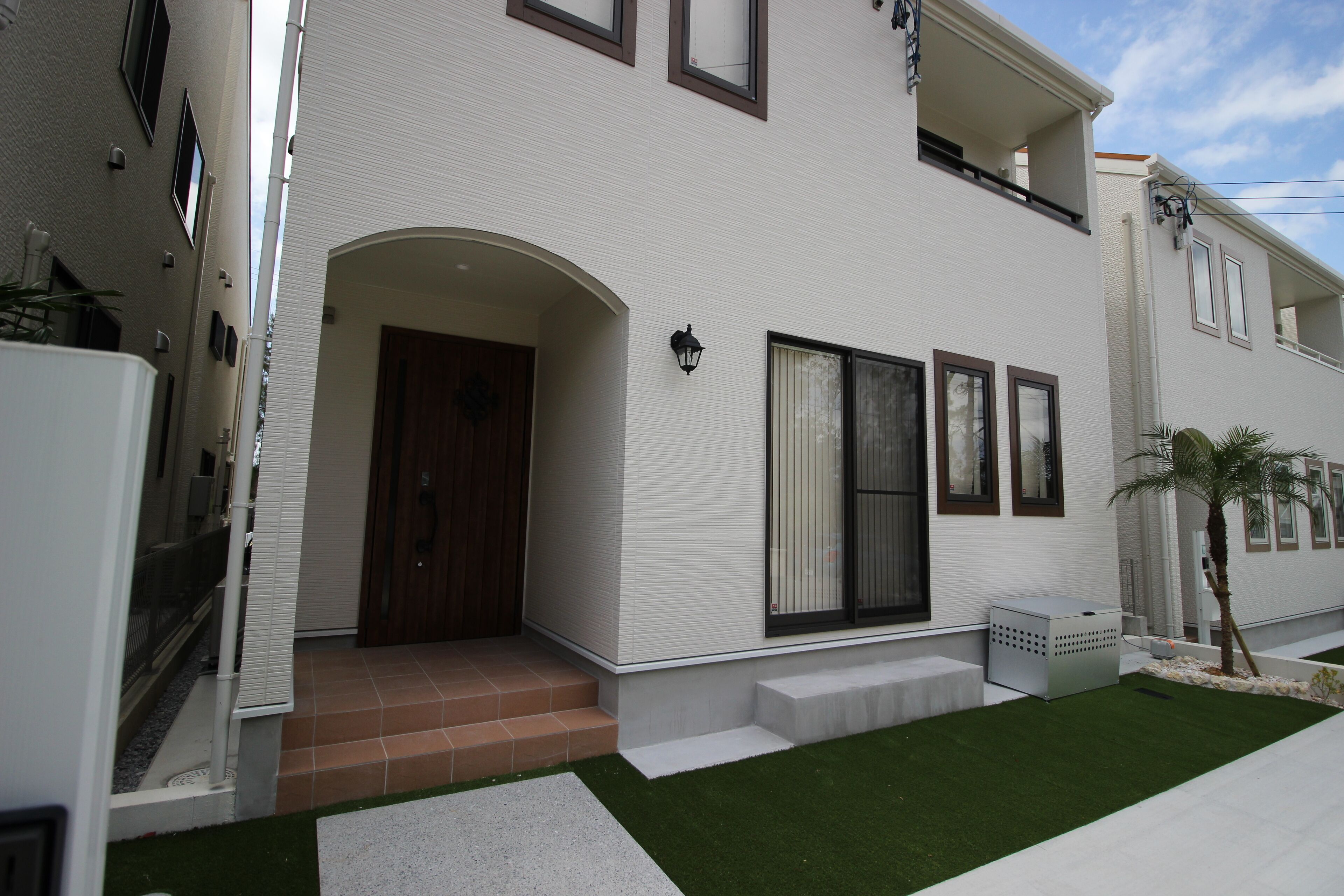 Edifici modern de dues plantes amb façana blanca, porta de fusta i un petit jardí davanter amb gespa artificial i una palmera.