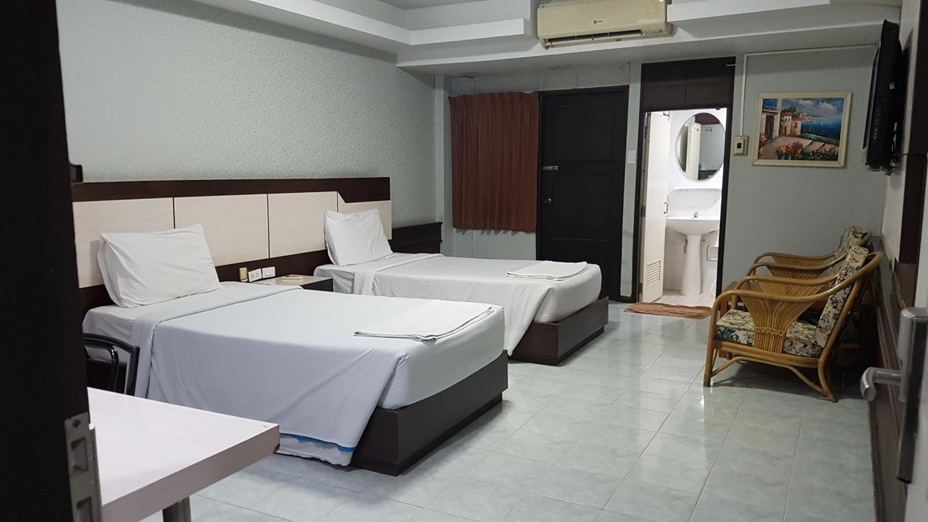 A hotel room with two beds, a small table, a chair, a bathroom with a sink, and a painting on the wall.
