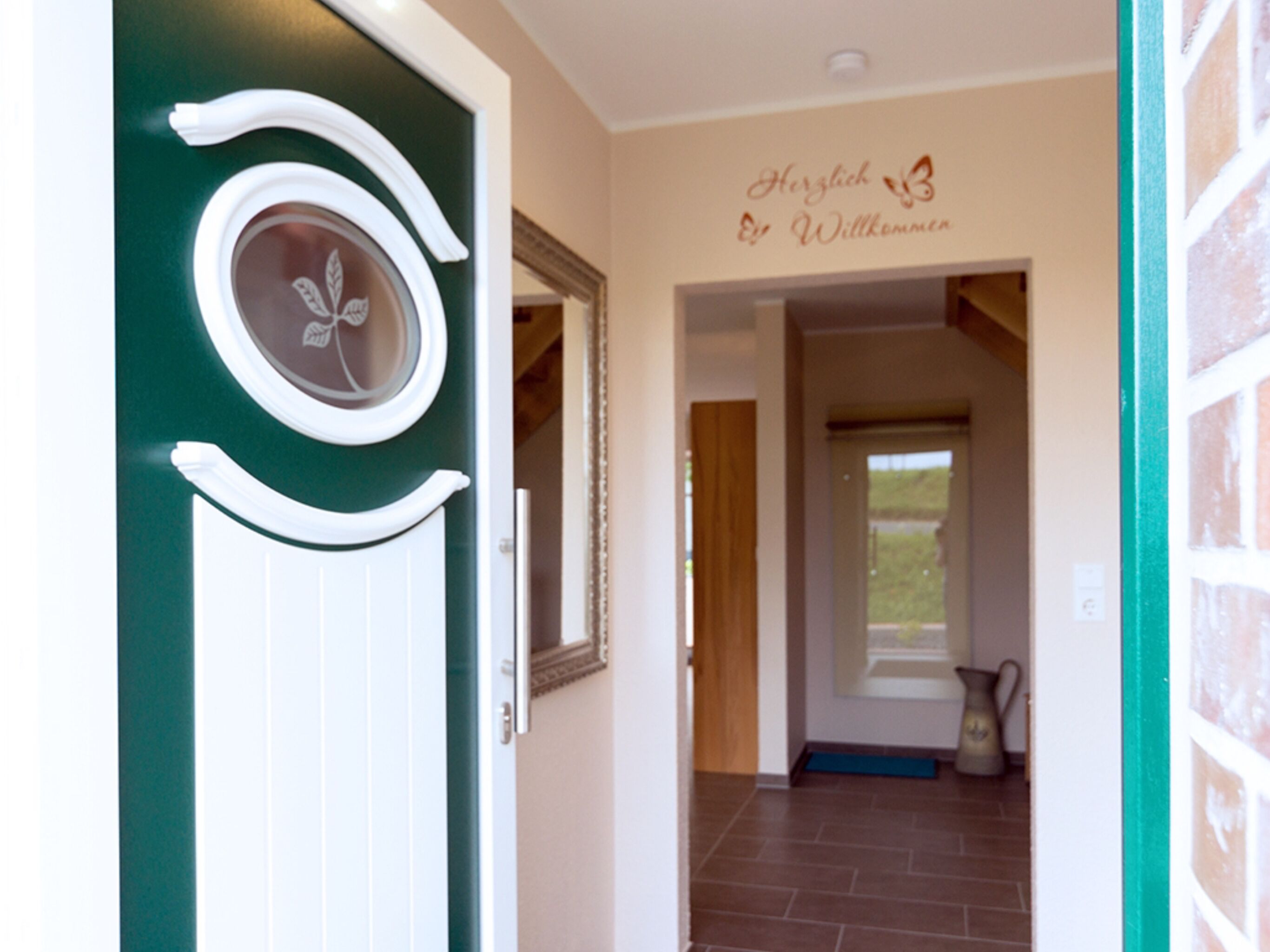 Ein Korridor mit einer grünen Tür, die ein rundes Fenster und einen dekorativen, weißen Rahmen hat. Auf der Wand ist ein Schild mit der Aufschrift 'Herzlich Willkommen'.