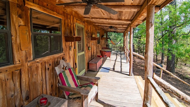 A wooden cabin with a porch, a ceiling fan, and a view of the forest.