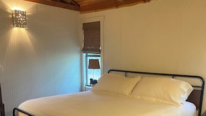 A bedroom with a wooden floor, a bed with white bedding, a patterned rug, a wall-mounted lamp, and a window with a blind.