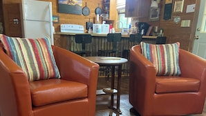 A cozy cabin interior with two leather armchairs, a wooden dining table, and a rustic cowhide rug.