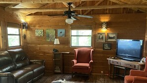 A cozy cabin interior with wooden walls, a leather sofa, a red armchair, a television, and a ceiling fan.
