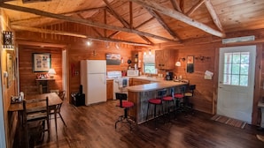 A rustic wooden cabin with a kitchen featuring a central island with bar stools, a dining area with a wooden table and chairs, and a white door.