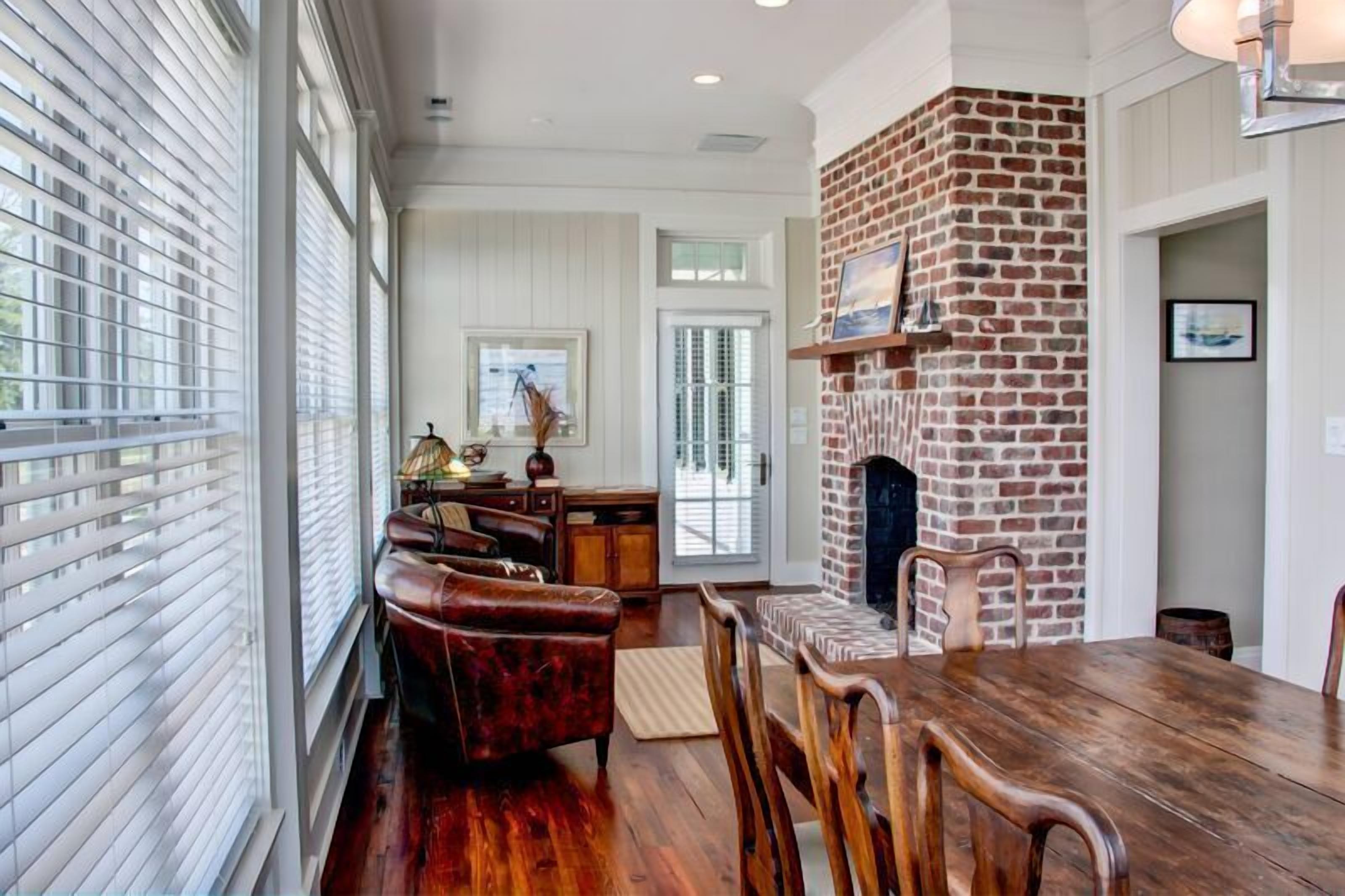 A dining area with a brick fireplace, wooden furniture, and large windows with blinds.